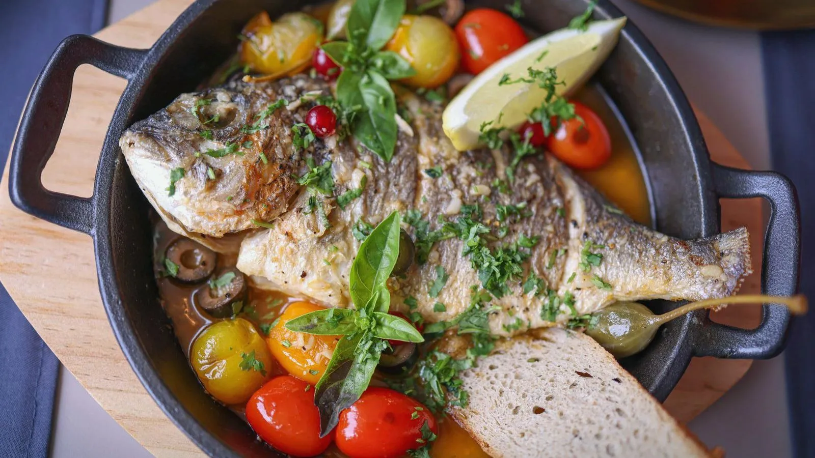 A whole baked fish with cherry tomatoes, olives, lemon and basil in a cast-iron pan, beside a slice of bread