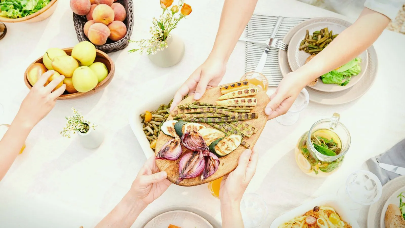 Overhead view of hands passing a board of grilled vegetables across a table set with salads, peaches and apples.