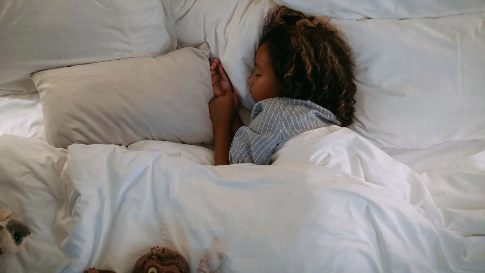 A young child asleep under a white duvet with soft toys beside the bed