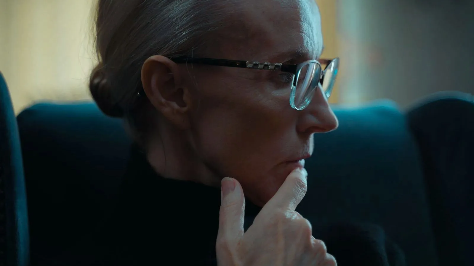 An older woman in profile wearing glasses, a hand resting against her chin