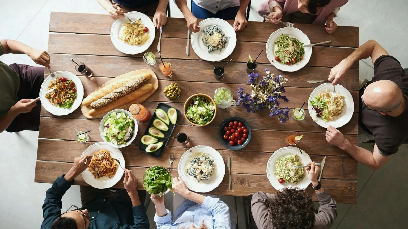 A group of people seated around a wooden table sharing plates of pasta, salad and bread, seen from above.