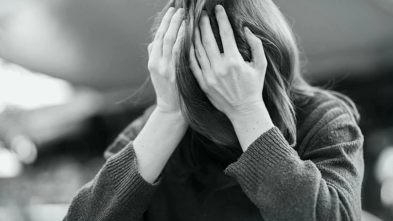 A black and white photo of a woman in a knitted sweater bending forward with both hands covering the head and face.