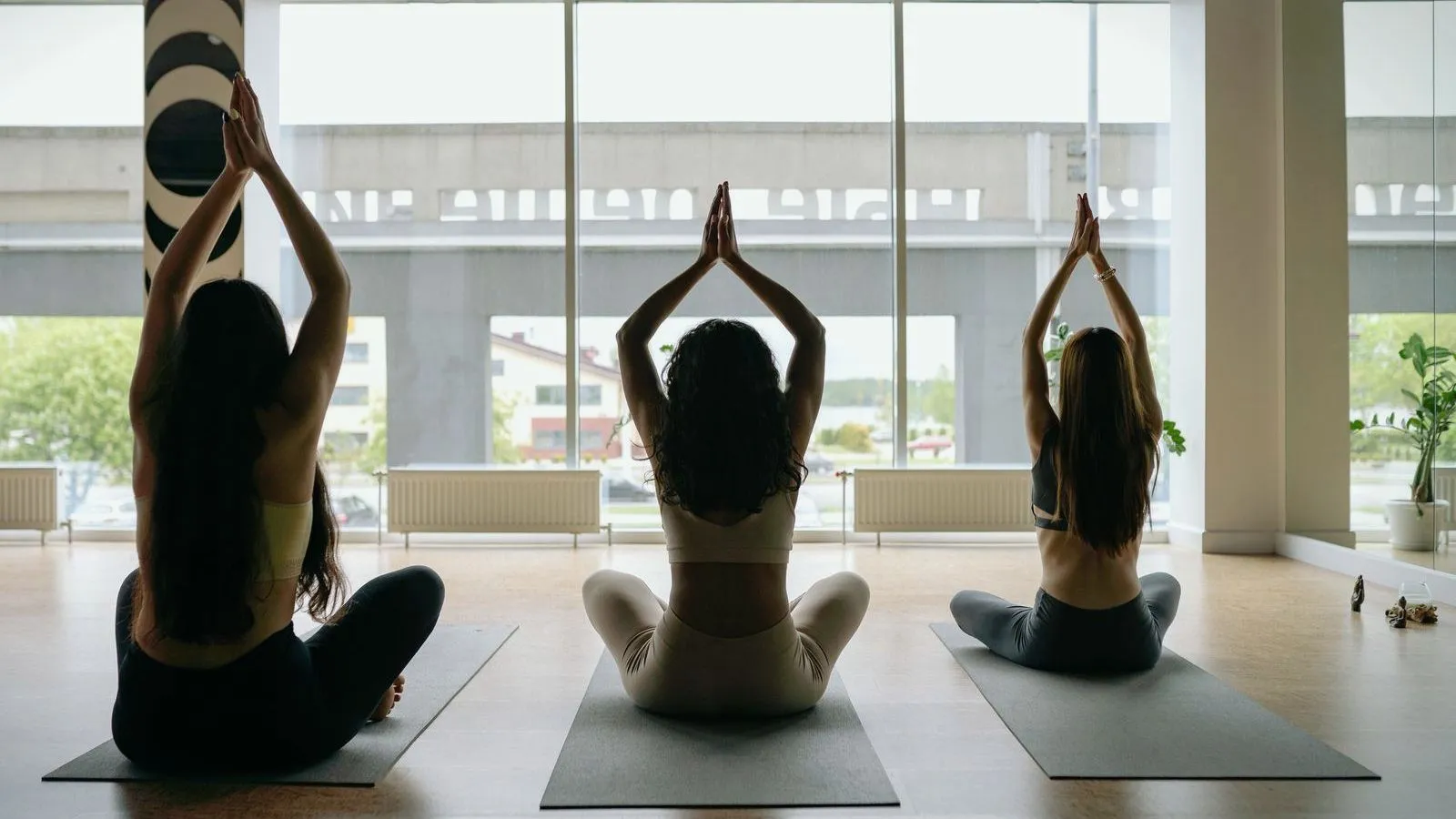 Three people seated cross-legged on mats with palms pressed together overhead, in a studio with tall windows