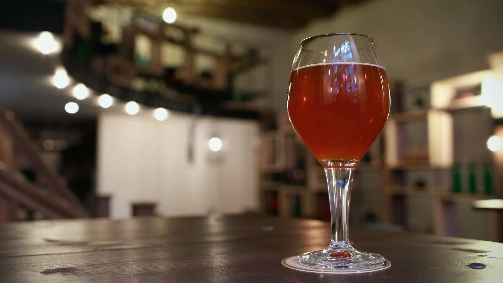 A single stemmed glass of amber beer on a wooden table in a dimly lit bar.