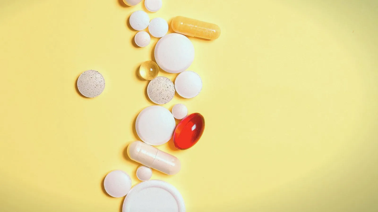 Assorted white tablets, capsules and a red softgel on a yellow background.
