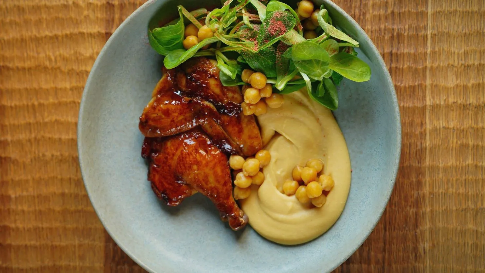 Glazed roast poultry beside a swirl of hummus topped with whole chickpeas and a handful of salad greens in a blue bowl