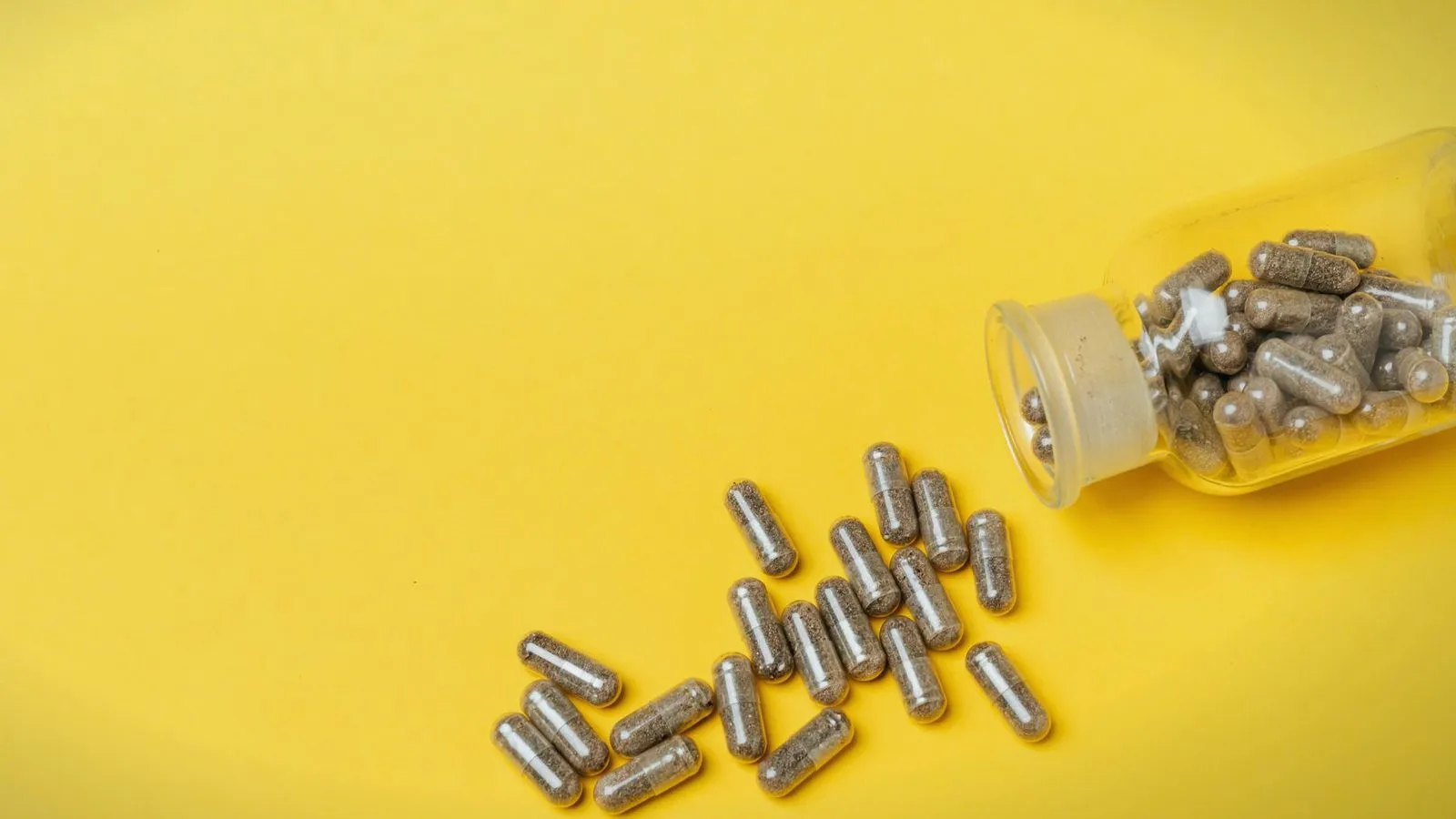 Grey capsules spilled from a clear bottle onto a yellow background