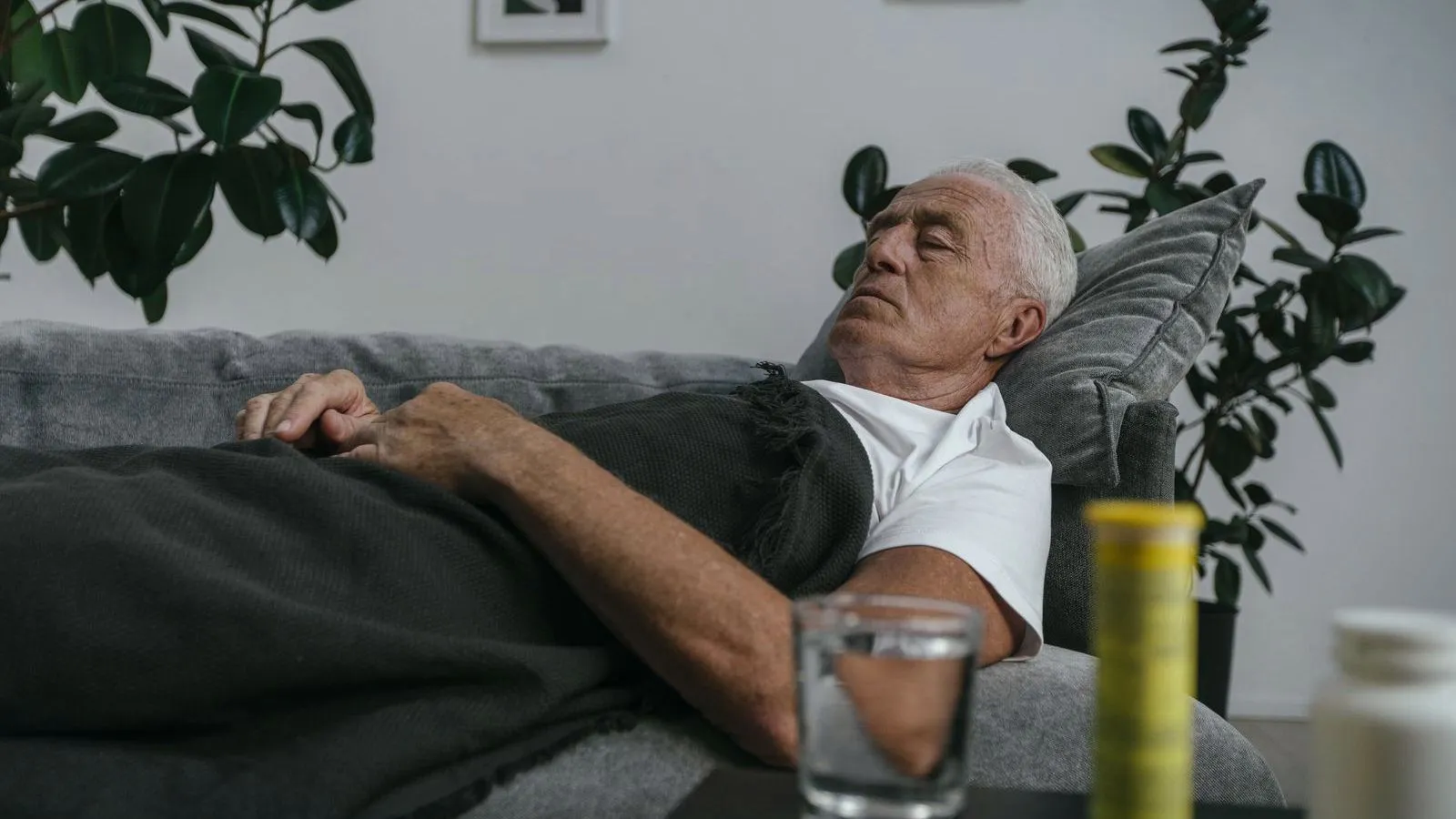 An older man napping on a sofa under a blanket, with a glass of water in the foreground.