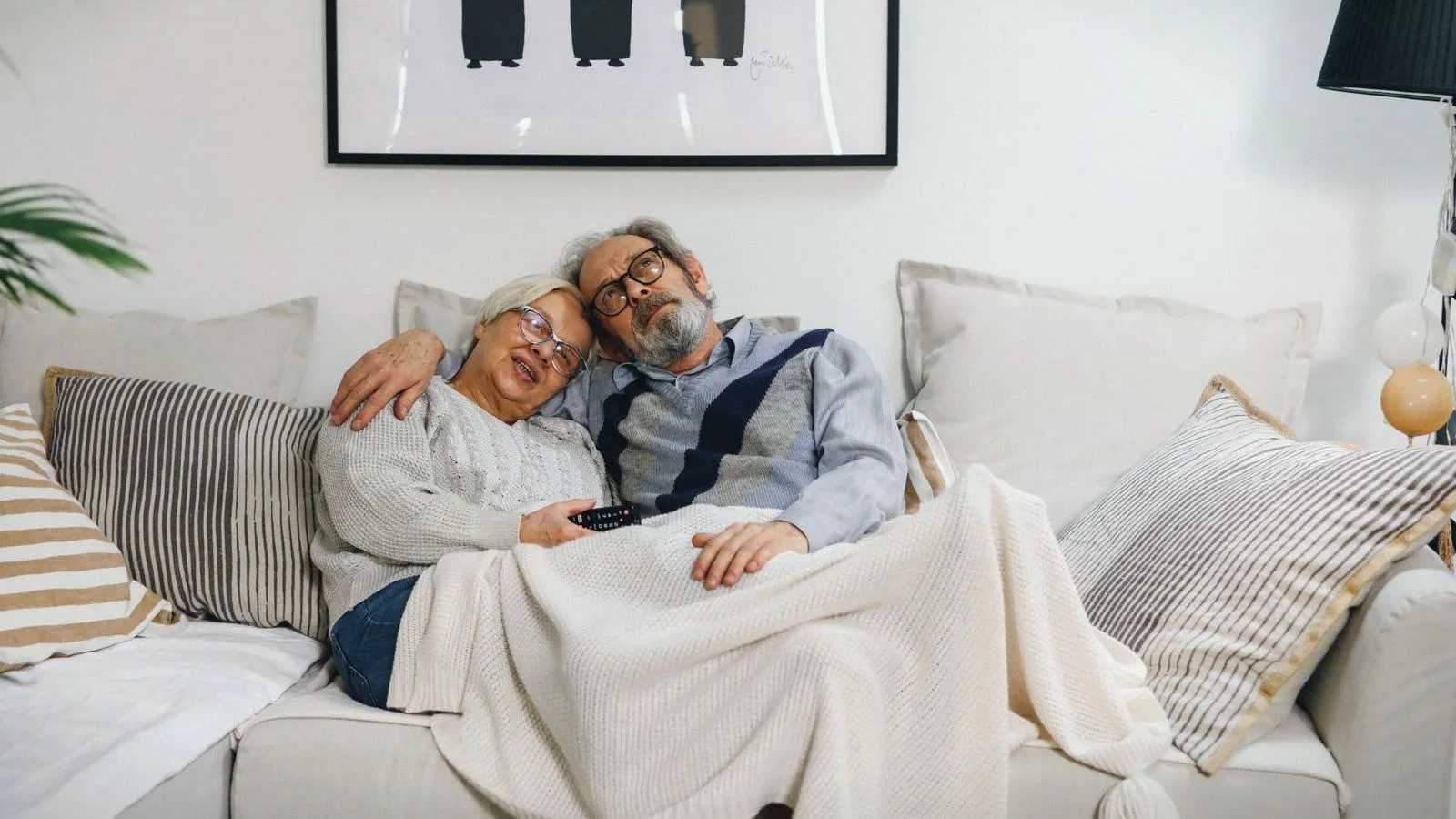 An older couple dozing on a sofa under a knitted blanket, a television remote still in hand