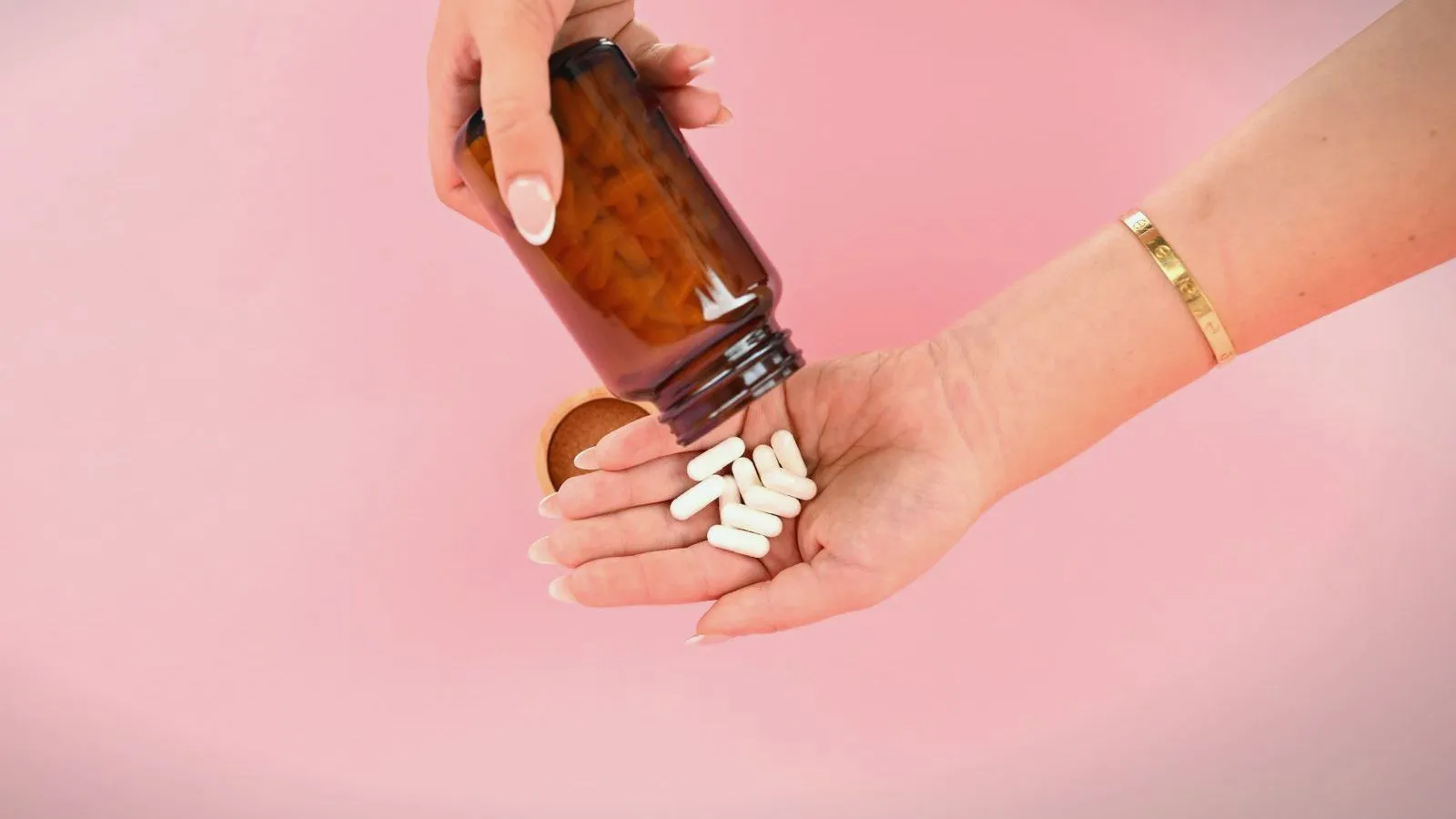 A hand tips white capsules from an unlabelled amber bottle into an open palm against a pink background.