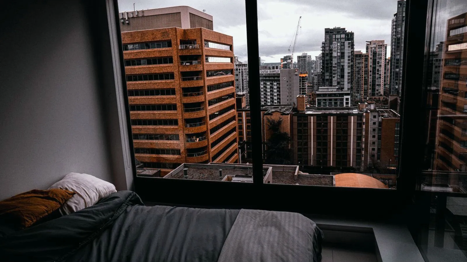 An unmade bed beside a large window looking out on lit city buildings
