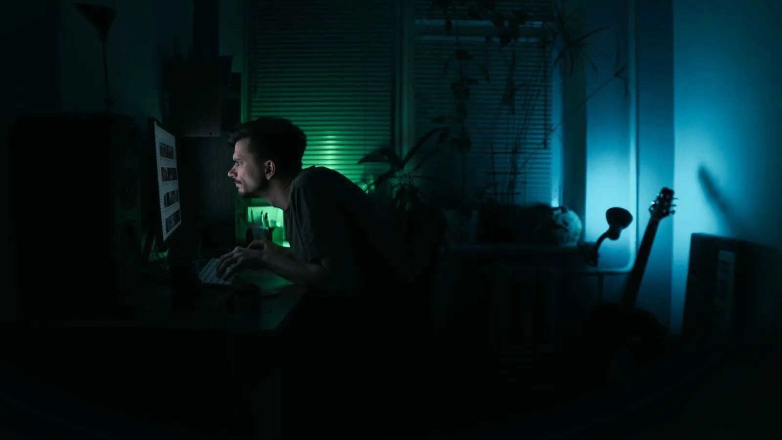 A person sitting at a computer in a dark room lit by green and blue light.