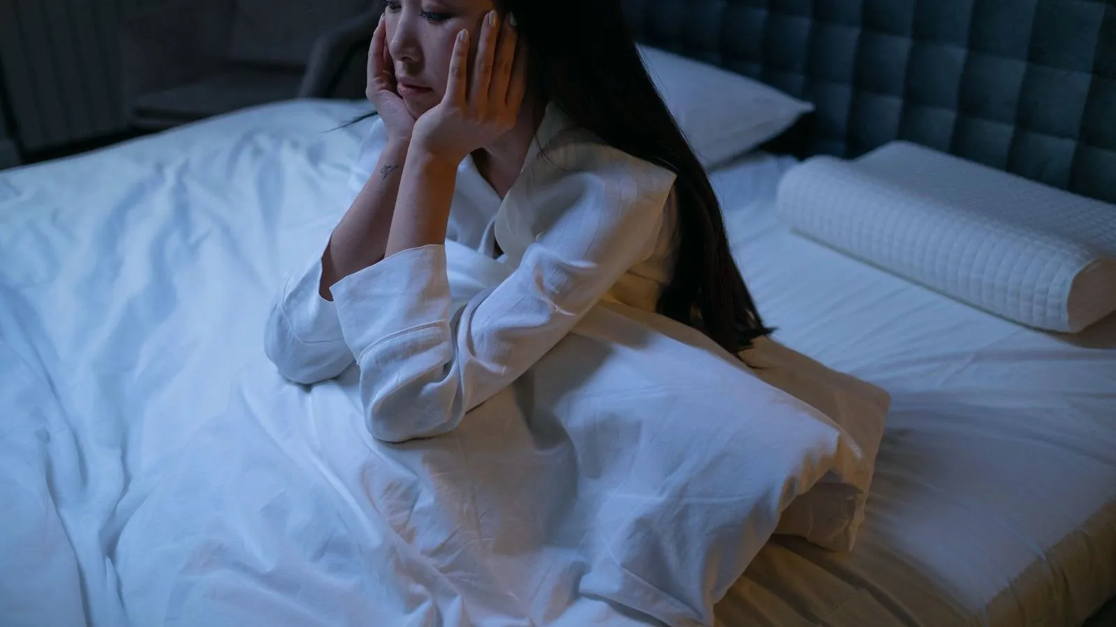 A person sitting up in bed at night in pajamas, head resting in their hands.