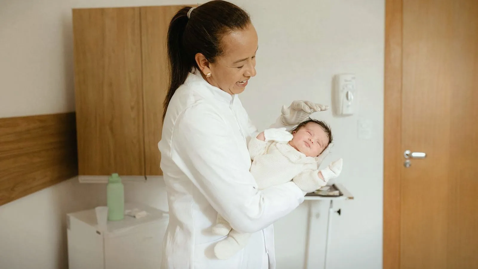 A health worker in white gloves holding a sleeping newborn in a clinic room