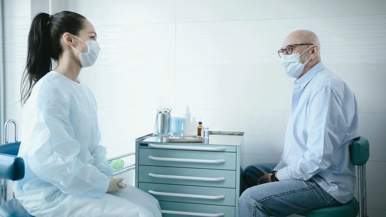 A masked clinician and an older masked patient sitting facing each other in a clinic room