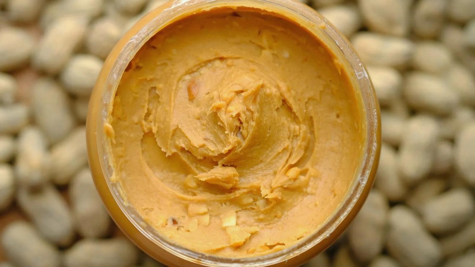 Overhead view of an open jar of peanut paste resting on a bed of shelled peanuts.