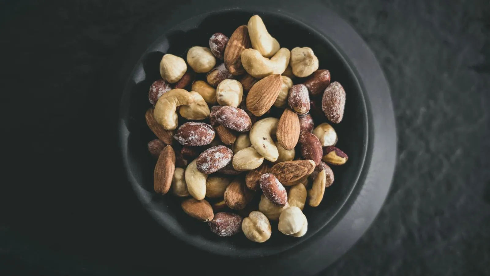 A dark bowl of mixed nuts, cashews, almonds, hazelnuts and peanuts, shot from above on a dark stone surface
