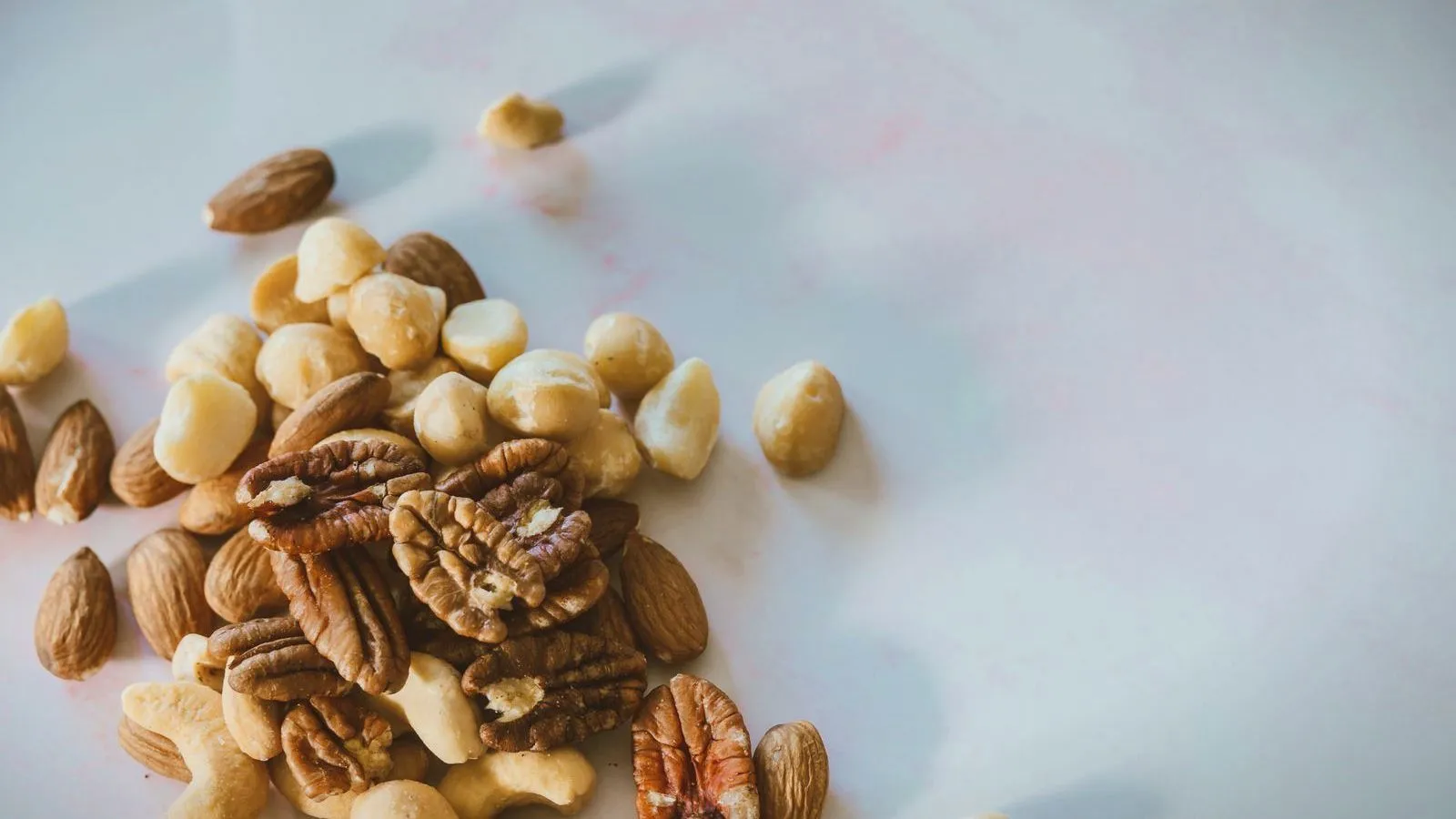 Almonds, pecans and other mixed nuts scattered on a pale surface.