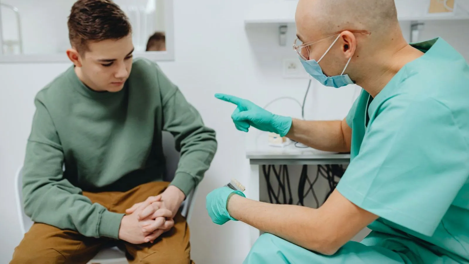 A teenager in a green sweatshirt sits with hands clasped while a clinician in teal scrubs, gloves and a face mask talks and gestures.