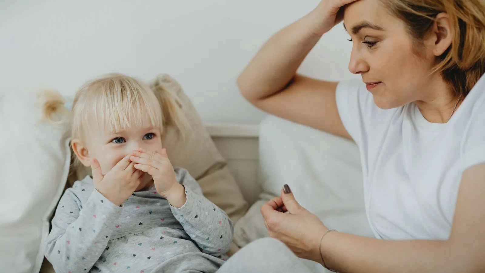 A toddler with both hands over the mouth sits on a sofa beside a watchful parent