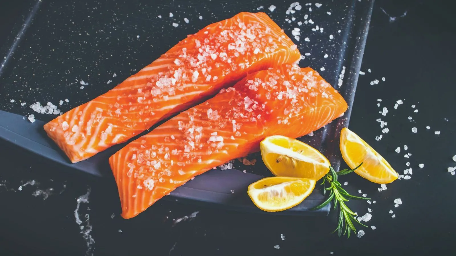 Two salted salmon fillets on dark slate with lemon wedges and rosemary