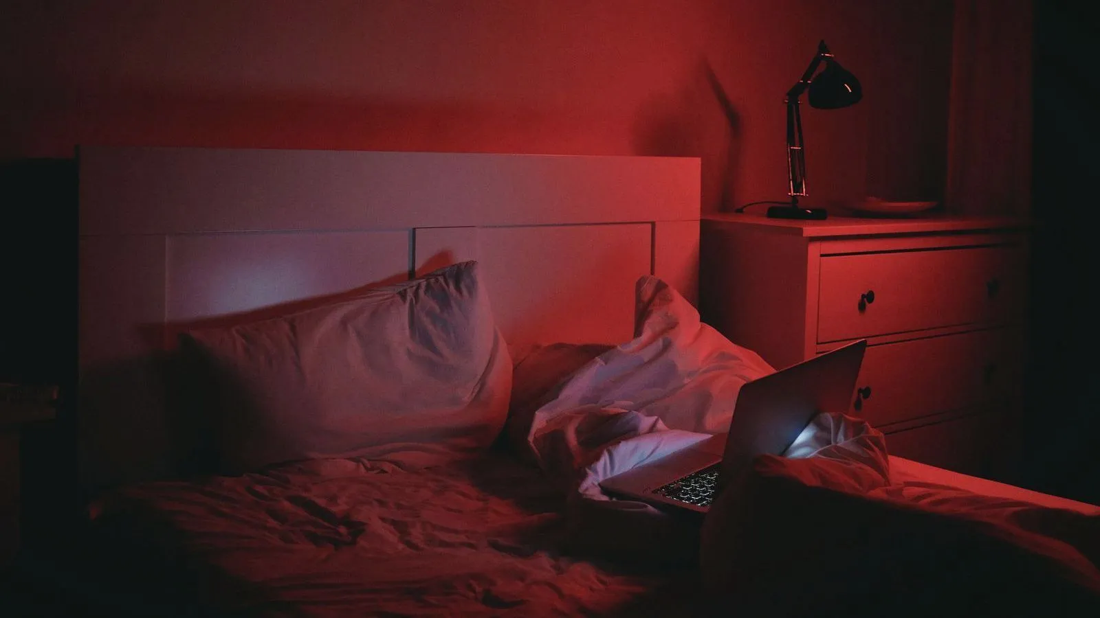 An open laptop glows on an unmade bed in a bedroom lit dim red, with a desk lamp on a chest of drawers beside it.
