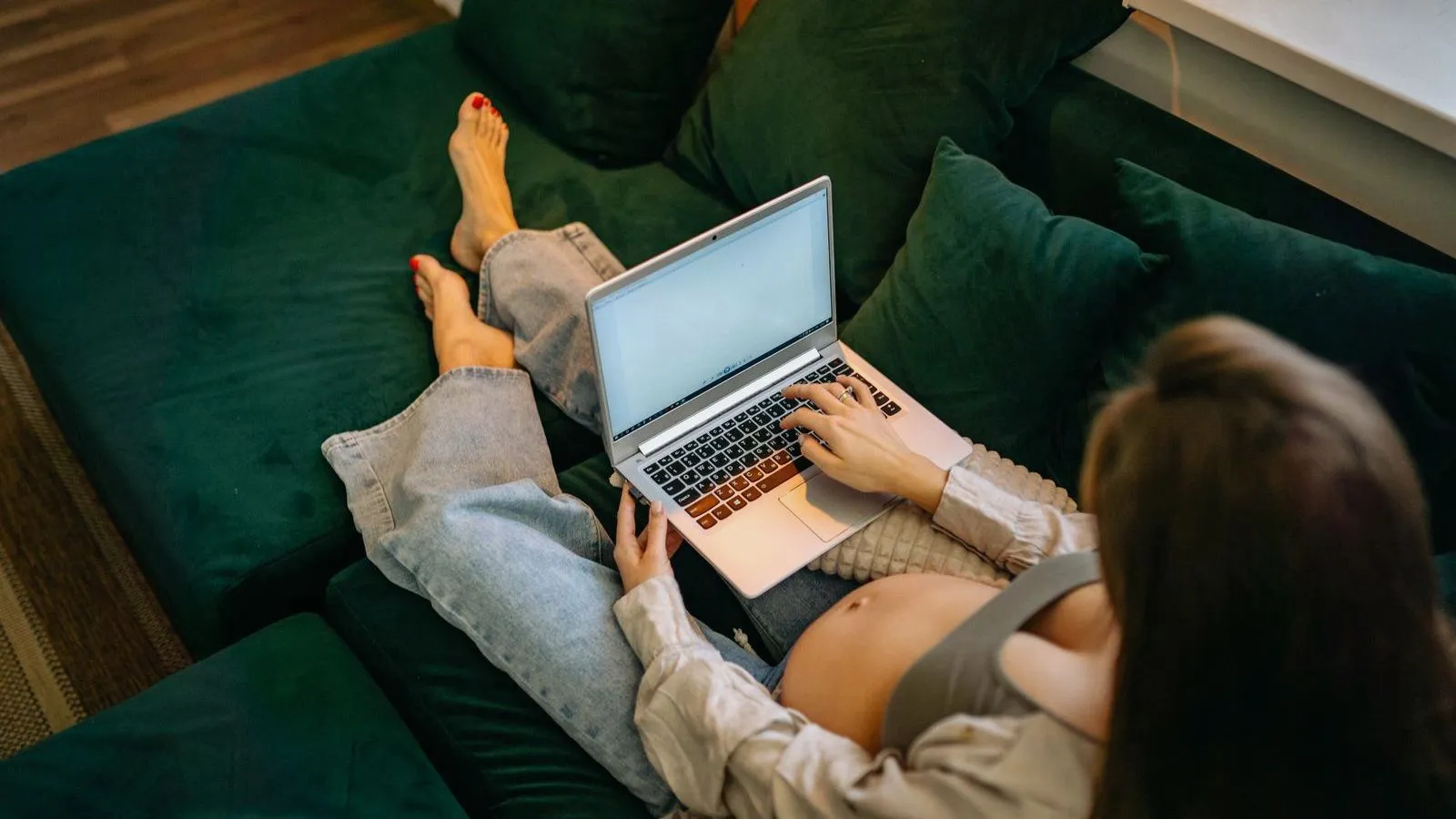 A pregnant person sitting on a sofa typing on a laptop, seen from above and behind.