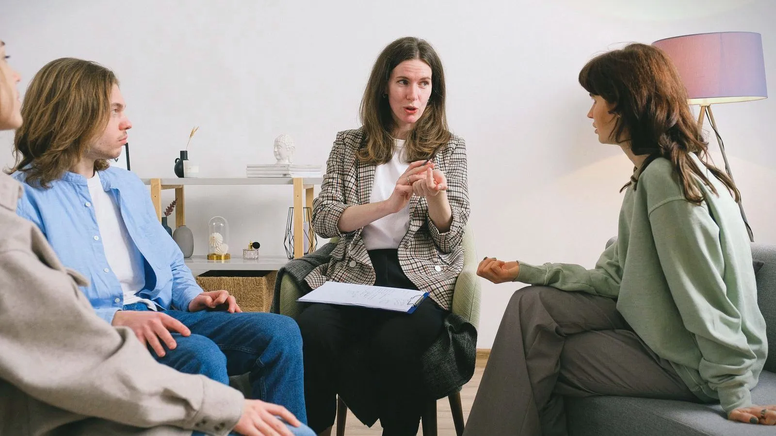 A small group sits in a circle in a living room while one of them, holding papers, speaks and gestures.