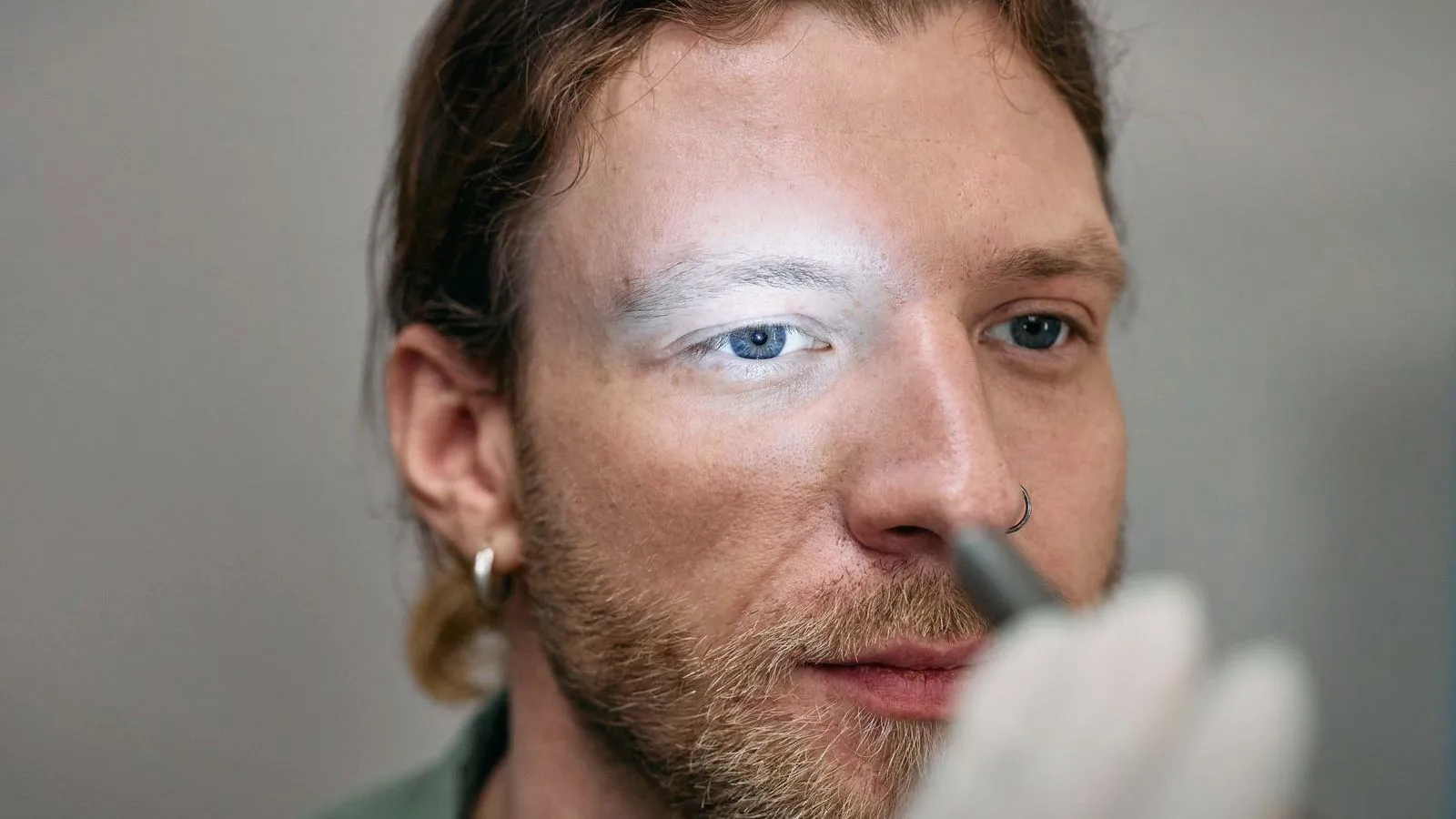 A gloved hand holds a small light up to the face of a bearded person, casting a bright patch across one eye and brow.