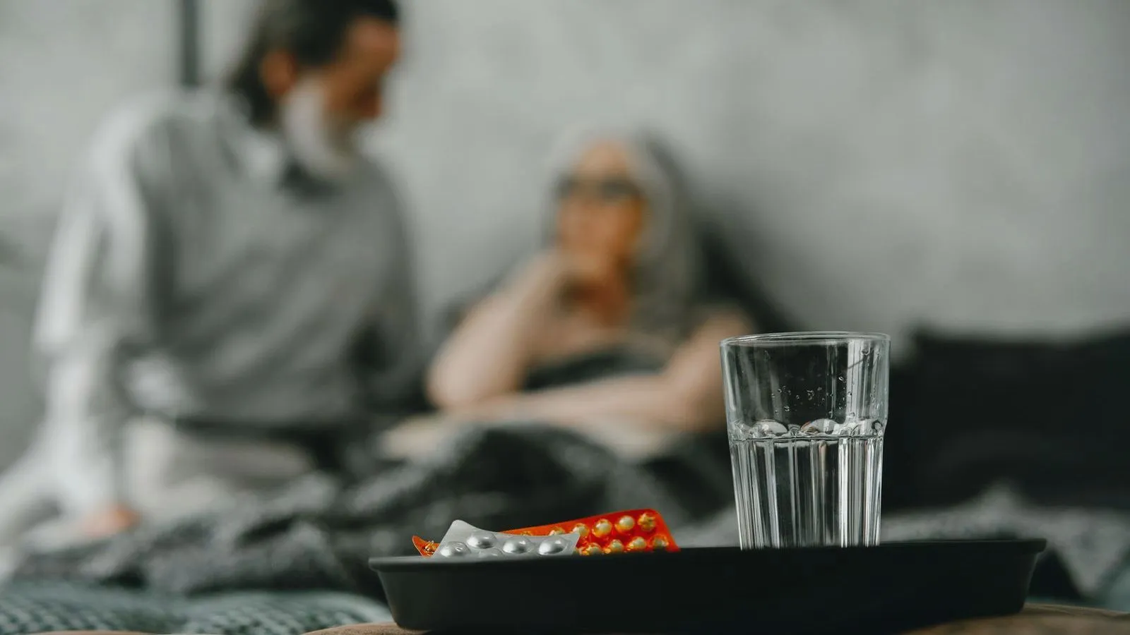 A glass of water and blister packs of tablets sit on a black tray, with an older man and woman blurred on a bed behind.
