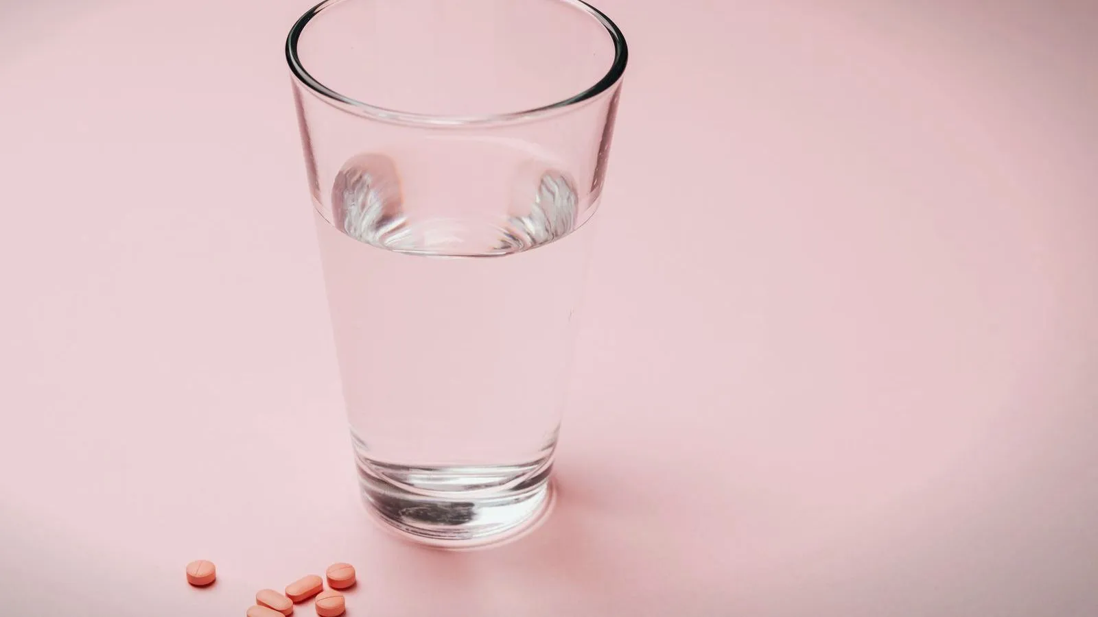 A glass of water with a few orange tablets beside it on a pink surface.