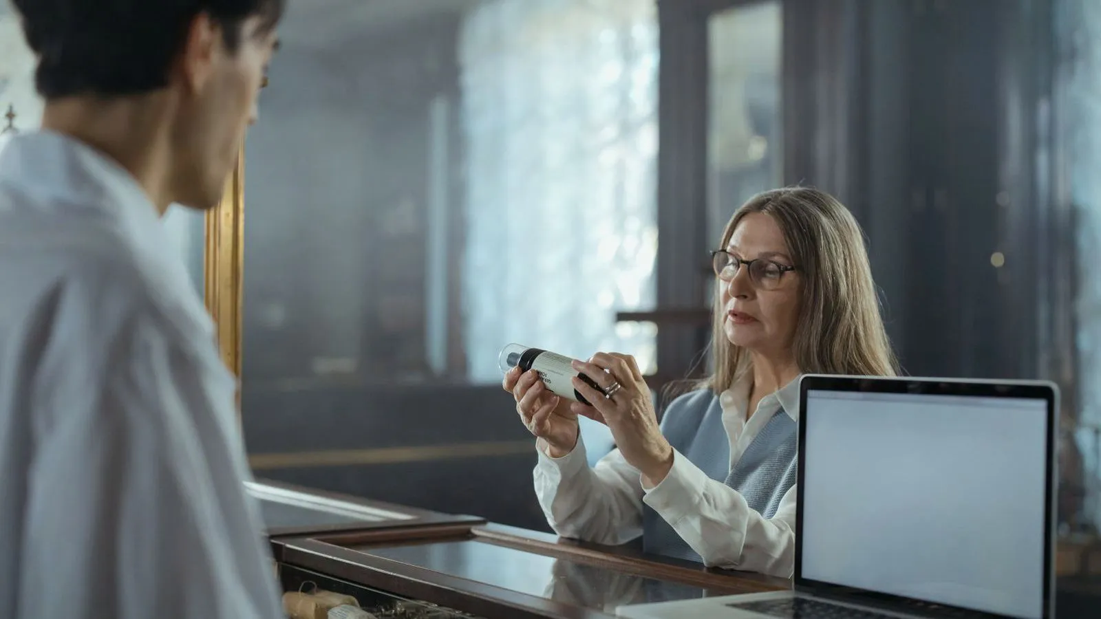 An older woman in glasses reads a small product at a shop counter beside a laptop while an assistant faces her.