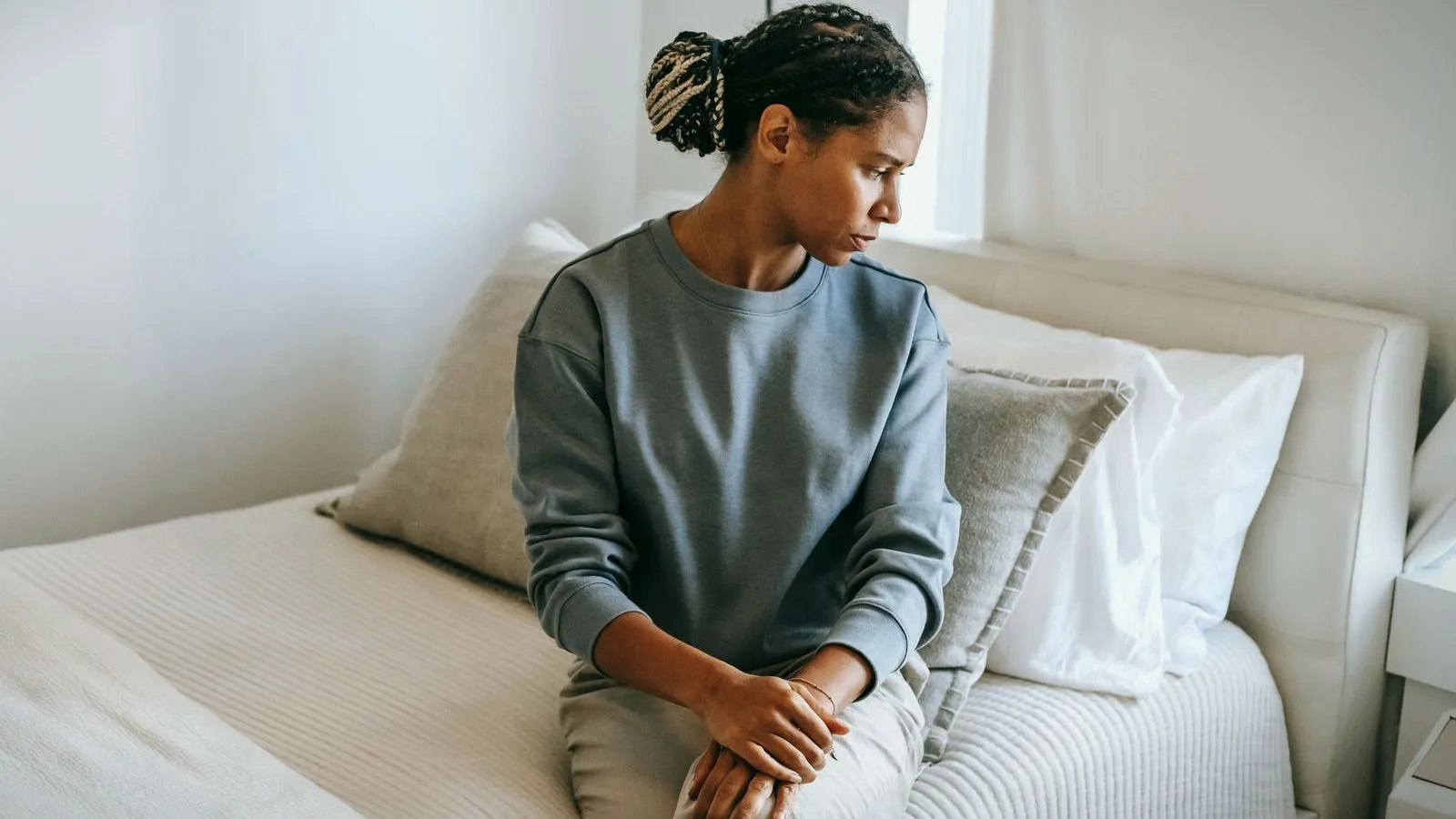 A young woman sitting on the edge of a bed with her hands clasped, looking down