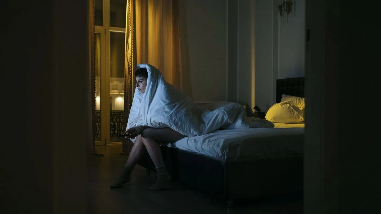 A person sitting on the edge of a bed at night wrapped in a duvet
