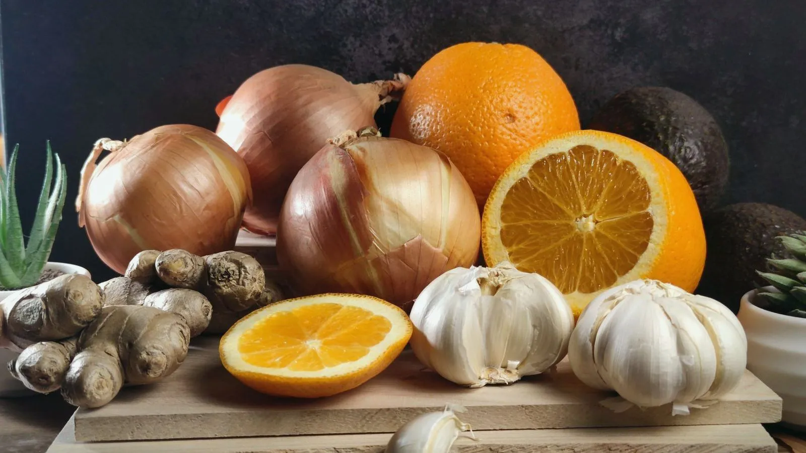 Onions, garlic, ginger and halved oranges arranged on a board against a dark background