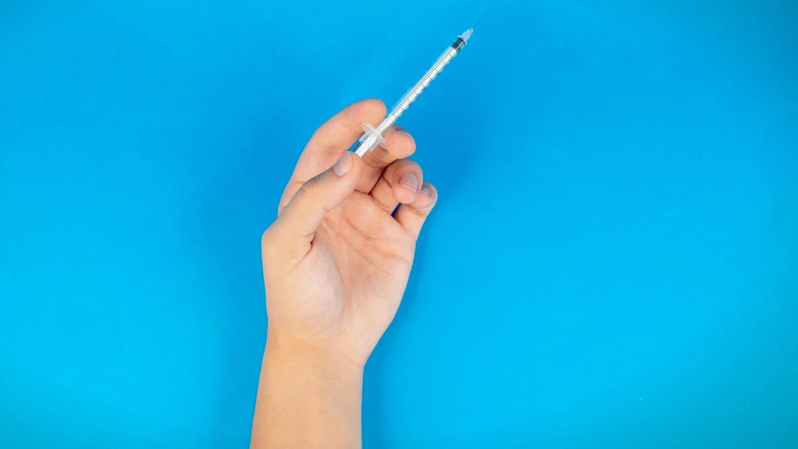 A hand holds up a thin syringe with a fine needle against a plain bright blue background.
