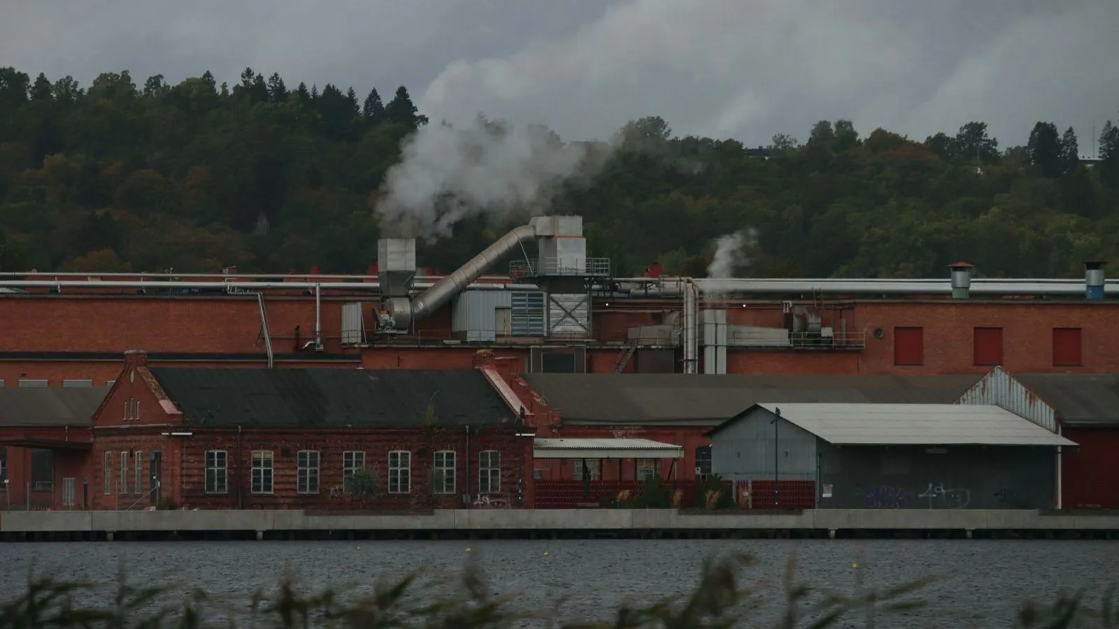 A red-brick factory on a riverbank with steam rising from its vents against wooded hills