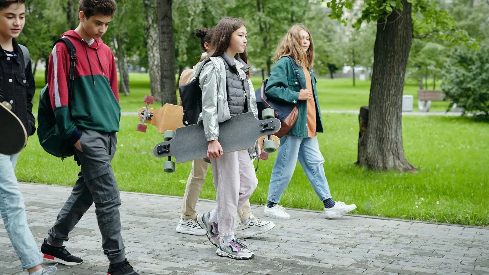 A group of teenagers walks along a paved path through a green park, two of them carrying skateboards.
