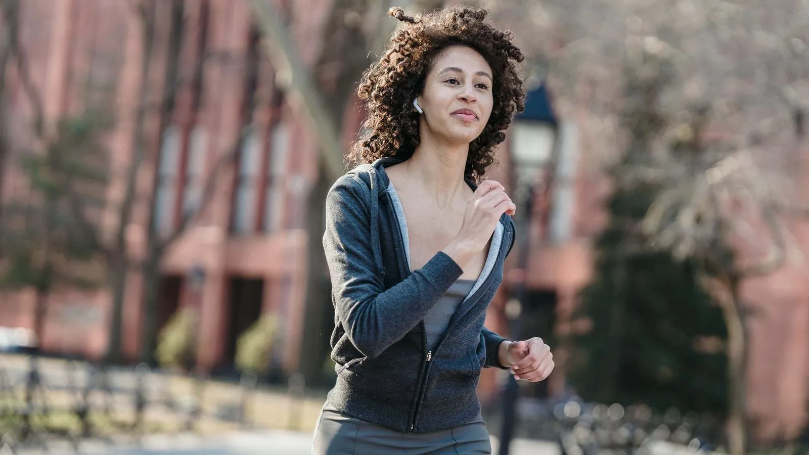 A woman in a grey hooded top jogging through a city park in early spring