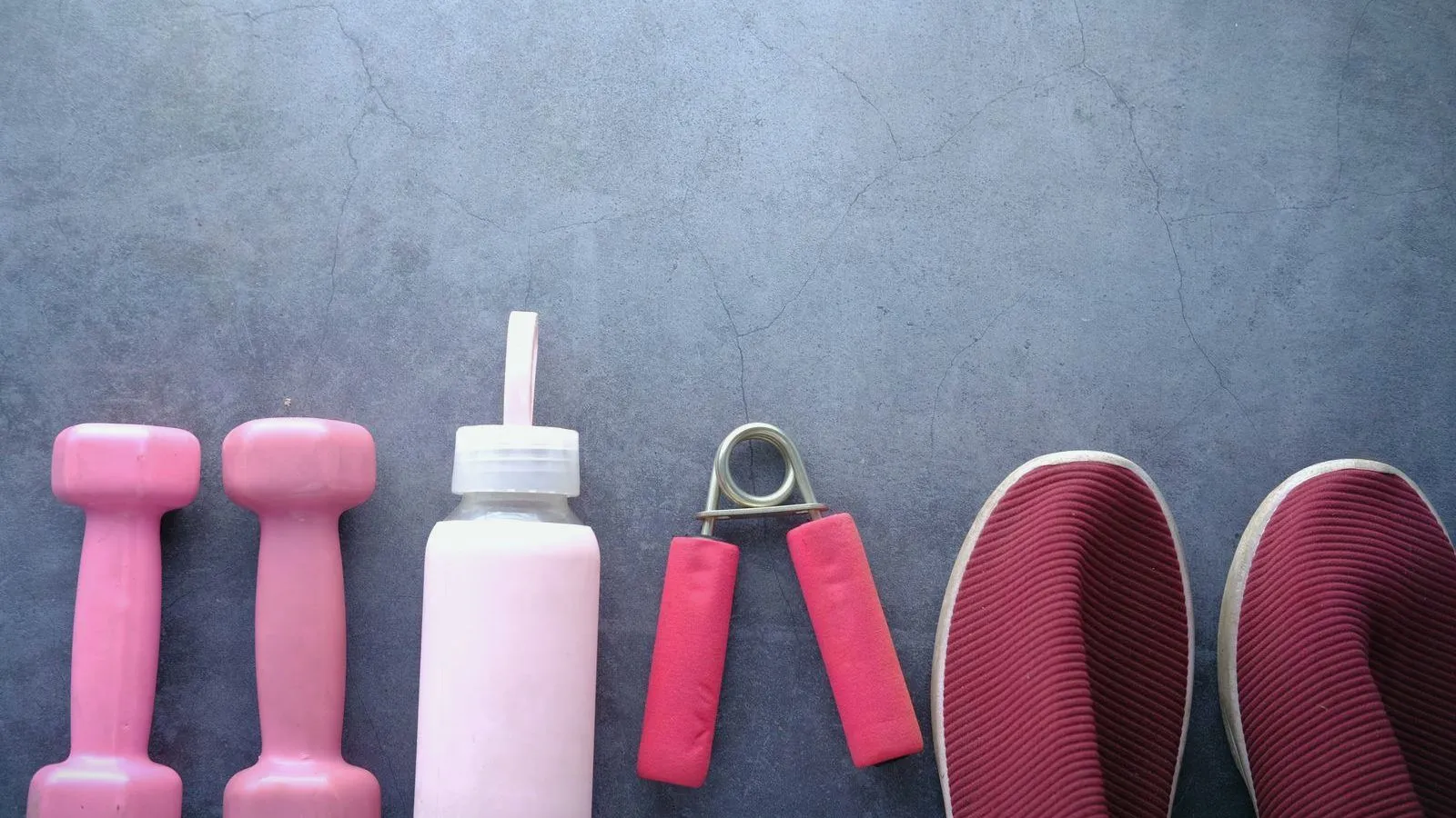 A row of pink dumbbells, a water bottle, a hand grip strengthener and a pair of shoes on a grey surface.