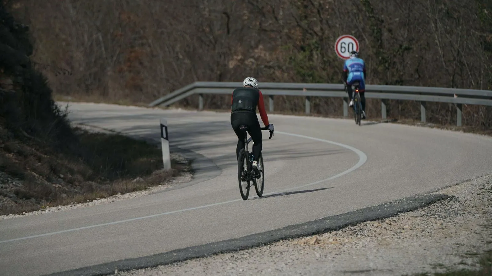 Two cyclists riding up a curving hill road beside a guardrail and a speed limit sign.
