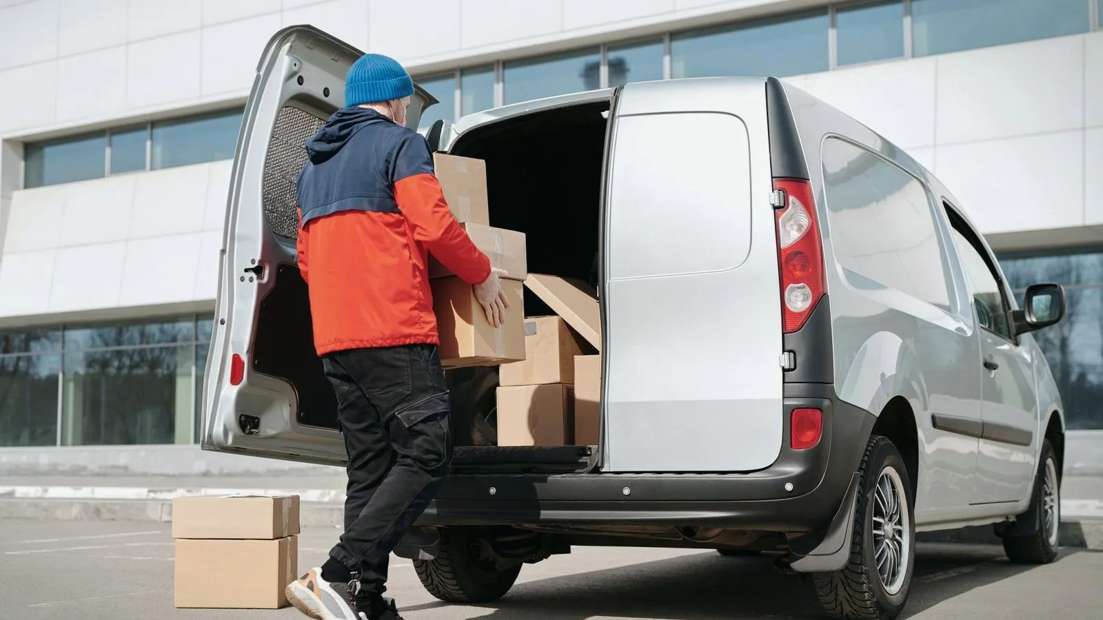 A worker in an orange and navy jacket and a blue hat, seen from behind, lifting cardboard boxes into the back of a silver van.