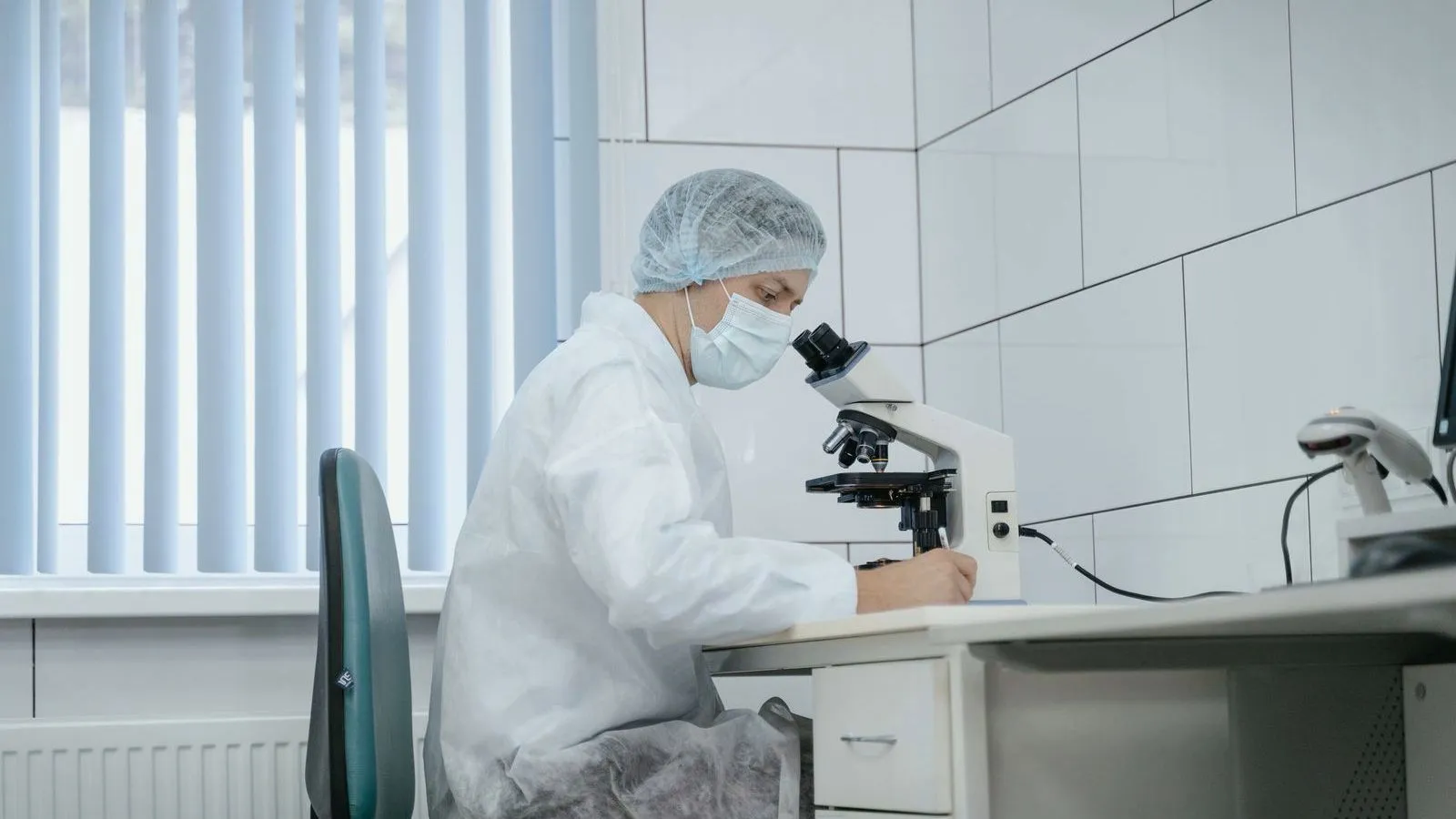 A laboratory worker in a white protective coat, hair cover and face mask writes notes at a bench beside a microscope.