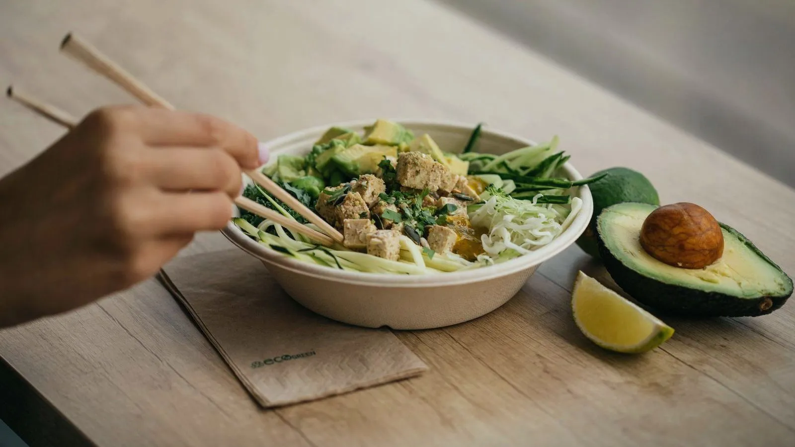 A hand holding chopsticks over a bowl of greens, noodles and tofu, with avocado and lime beside it