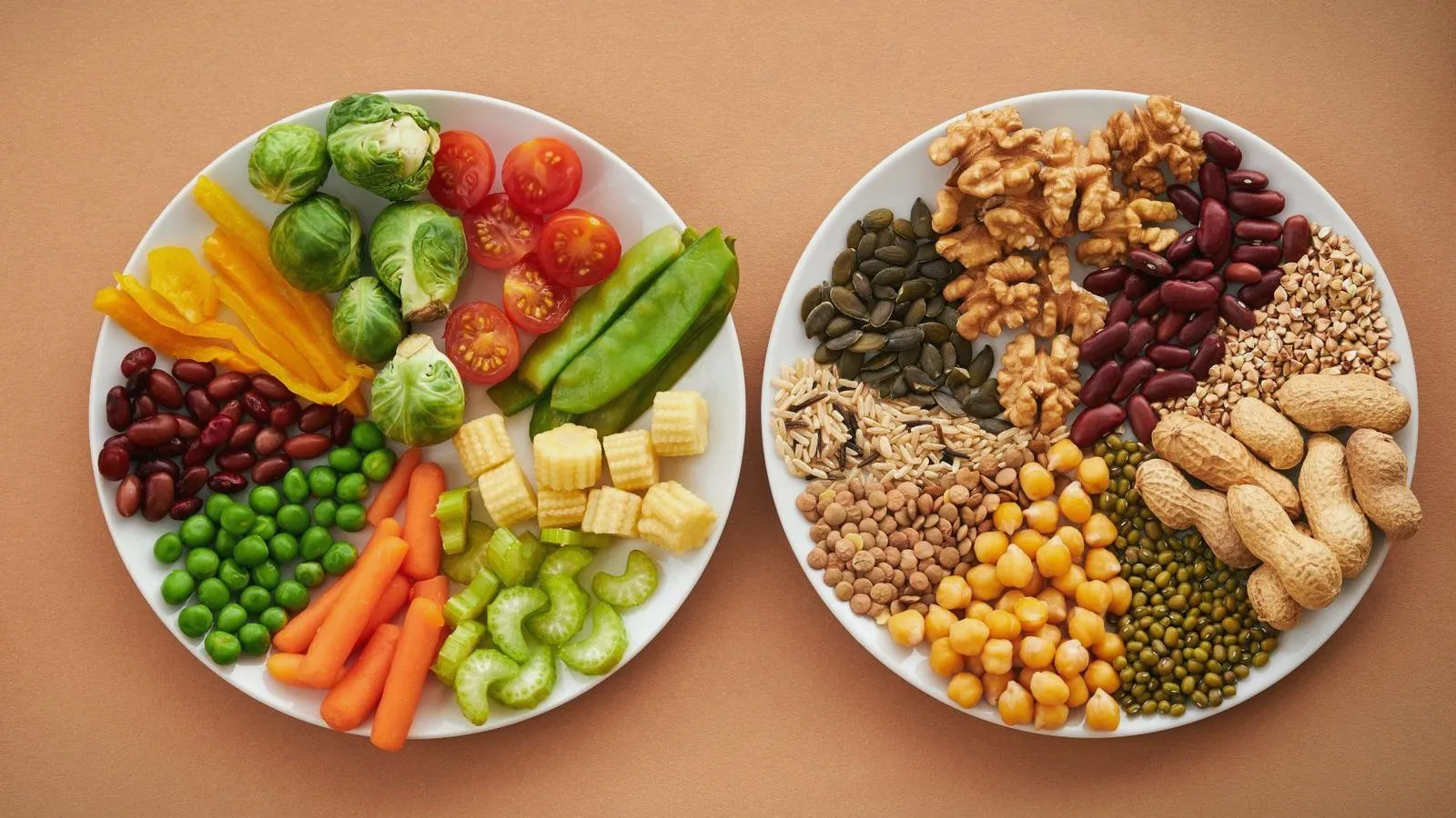 Two white plates seen from above on an orange surface, one of raw vegetables and one of nuts, beans, seeds and grains.