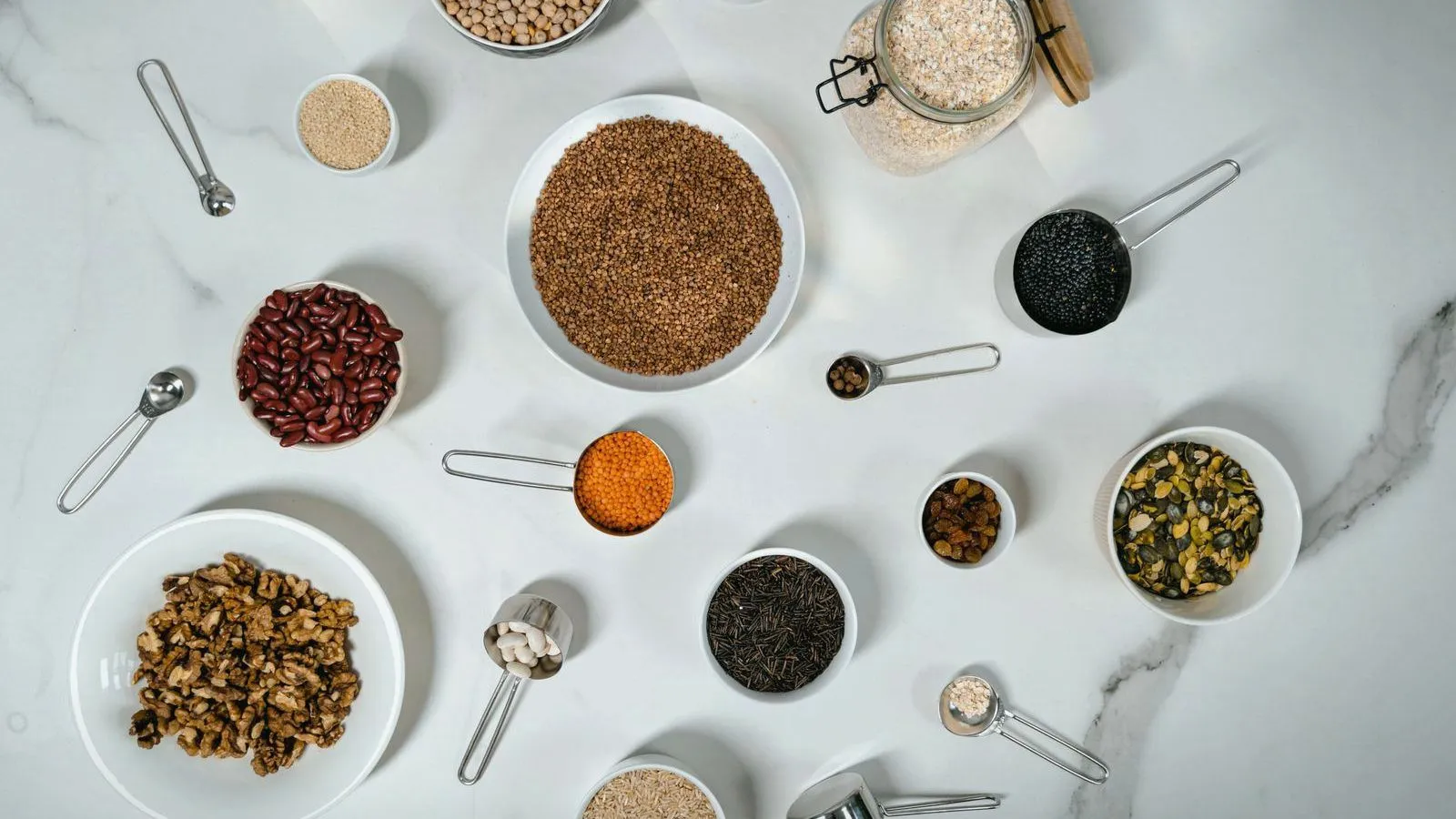 An overhead arrangement of small bowls and measuring spoons holding beans, lentils, grains, seeds and walnuts