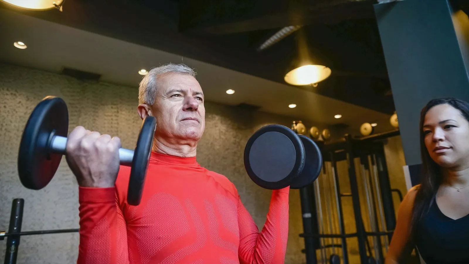 An older man in a red top pressing dumbbells overhead beside a trainer