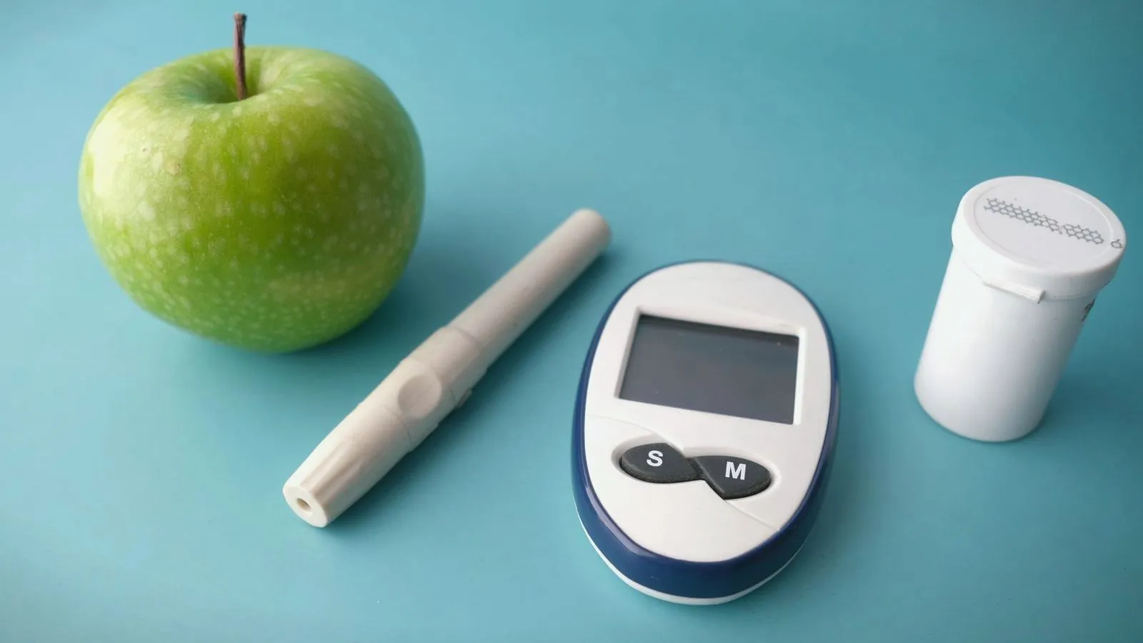 A blood glucose meter, a lancing pen and a test strip pot beside a green apple on a teal surface.