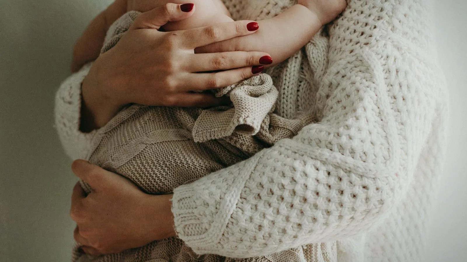 Close-up of a parent in a cream knitted sweater holding a baby wrapped in a knitted blanket, faces out of frame.