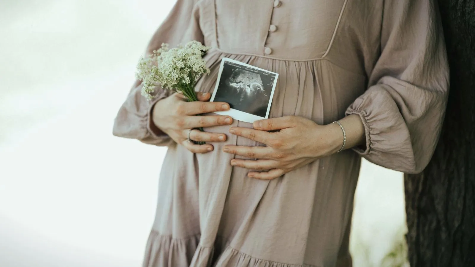 A pregnant person in a long dress holding a small bunch of flowers and an ultrasound print against their belly.